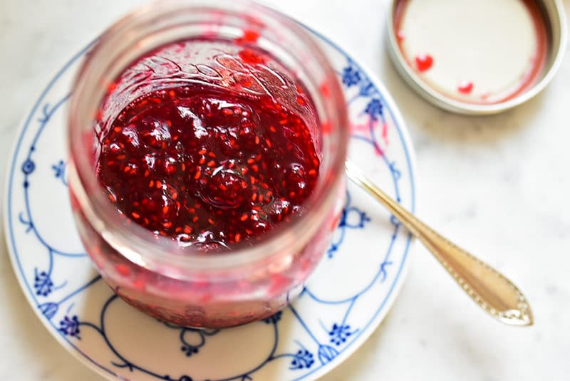 Overhead shot of a jar of raspberry jam on a blue and white plate with a spoon.