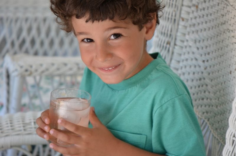 Little John with a glass of strawberry rose lemonade, sitting on a white wicker chair.