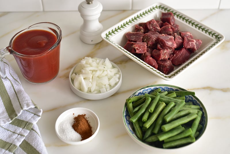 Ingredients on the counter with tomato sauce, salt and cinnamon, beef pieces, diced onion, and green beans