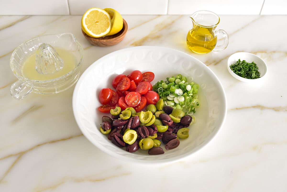 A white bowl with separate piles of tomatoes, scallions, and olives with a cut lemon on the side