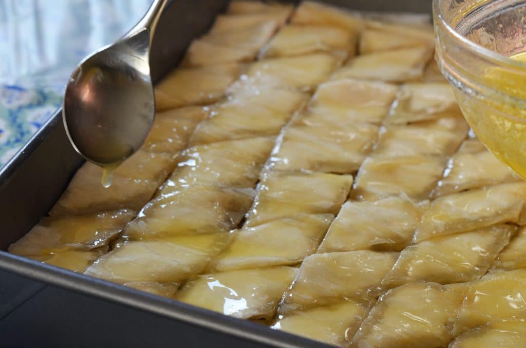 A spoon hovers over a pan of diamond cut baklava, drizzling butter over the top