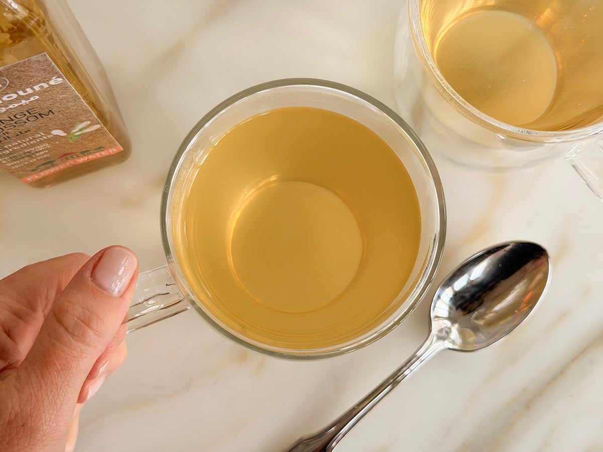 A hand holding a clear mug of cafe blanc with a spoon on the side