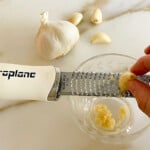 a clove of garlic rubbed over a microplane grater with two fingers holding the garlic, over a glass dish