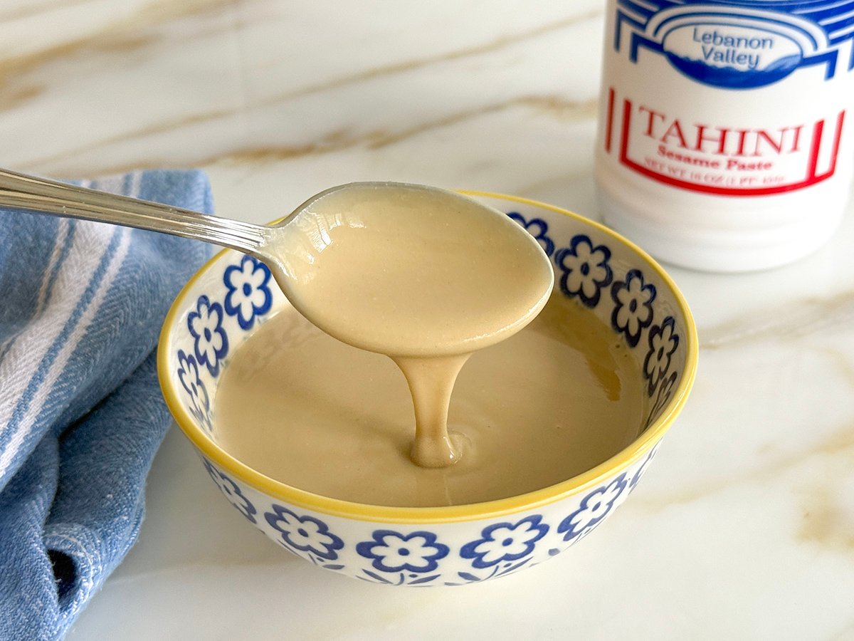 A spoonful of tahini over a blue and yellow bowl with a jar of tahini in the background