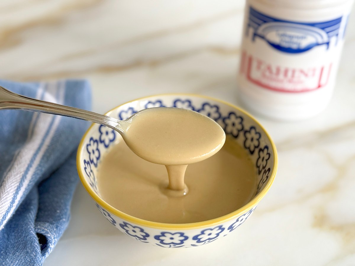 Tahini in a bowl with a spoon drizzling it in and a jar in the background