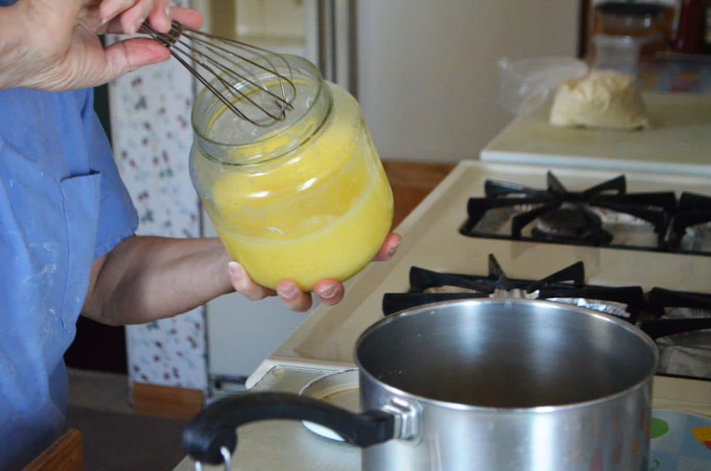 Two hands hold a jar of clarified butter, taking some out with a whisk over a pot on the stove
