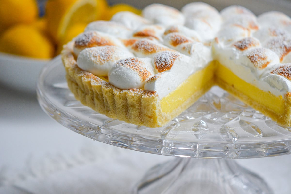 Lemon meringue tart on a glass pedestal plate with a slice cut out and lemons in the background