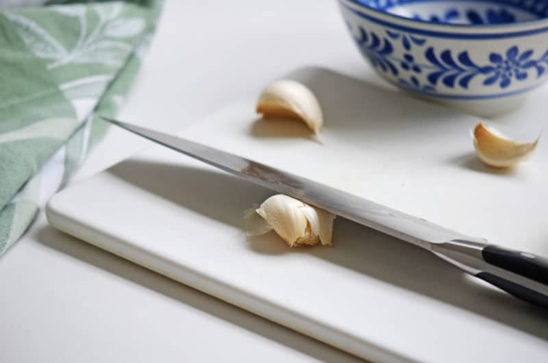 garlic under a large chefs knife with a blue bowl and green towel off to the side