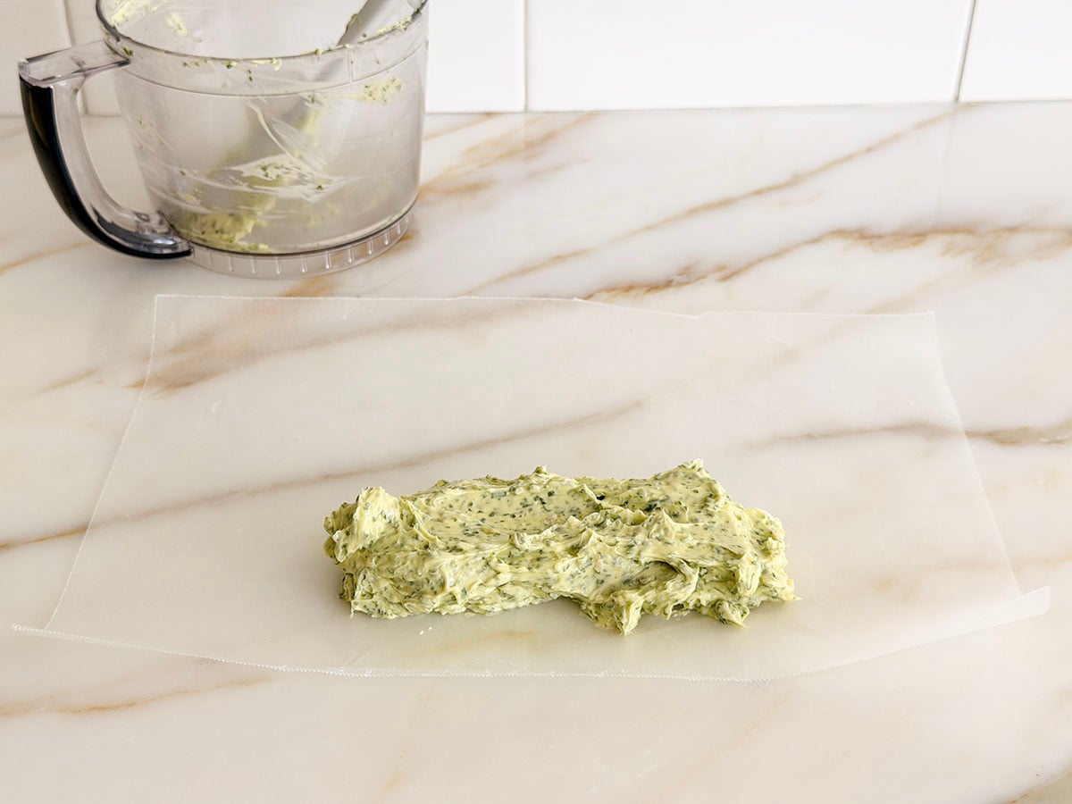 Very soft butter flecked with herbs spread into a log shape on a piece of wax paper