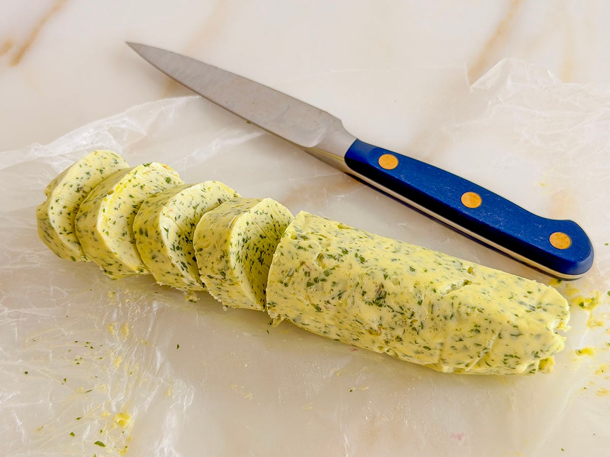 A log of compound butter with half of it cut in round slices with a blue knife on the side