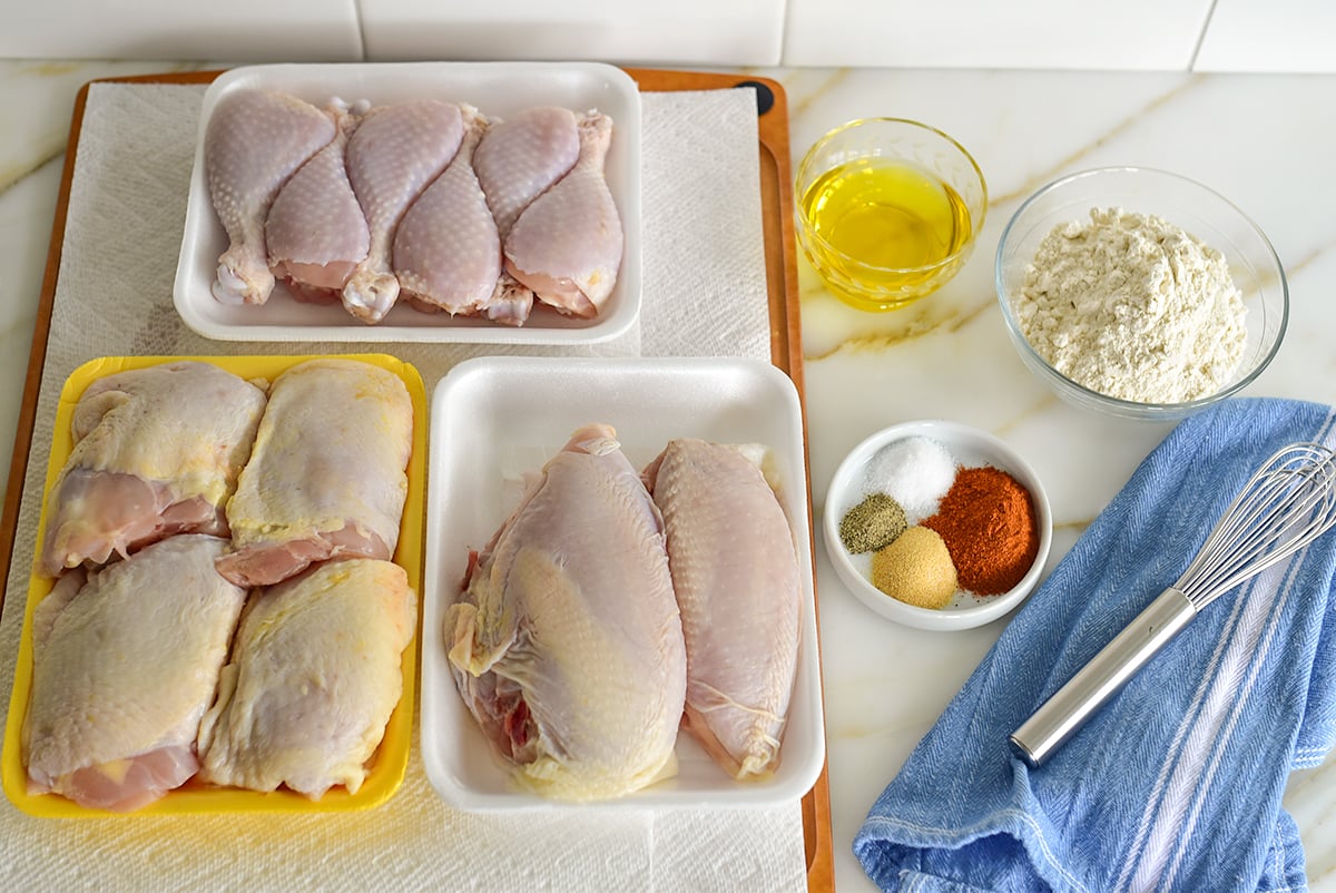 Ingredients for oven fried chicken on the counter in small bowls with a blue towel and whisk