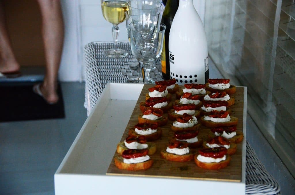 A wicker sideboard with za'atar roasted tomatoes and labneh on crostini, served on a cutting board and surrounded by champagne glasses, Maureen Abood