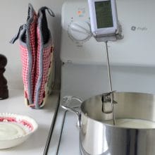 A pot with a thermometer in it on the stove with a little bowl of yogurt starter to make homemade yogurt