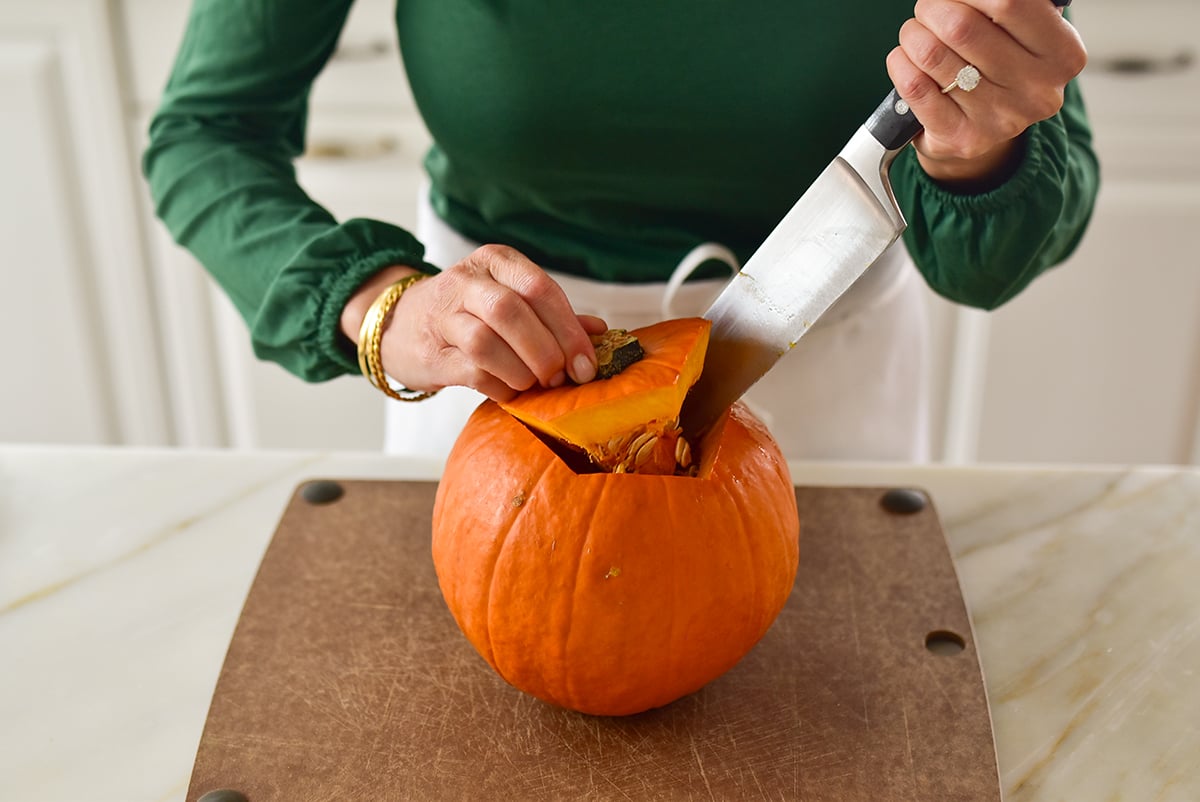 Two hands with green sleeves holding a chef's knife cutting the top off a pumpkin