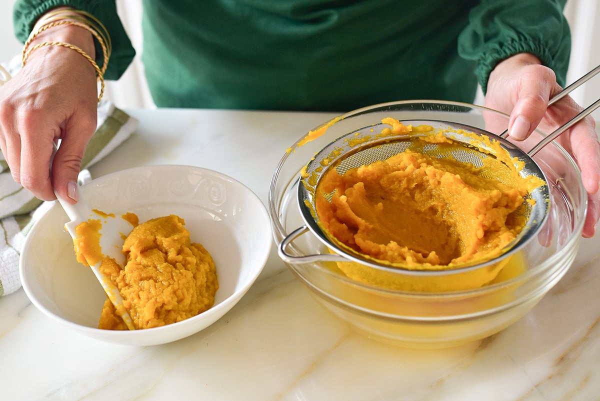 Two hands transferring pumpkin puree from a fine mesh strainer to a white bowl