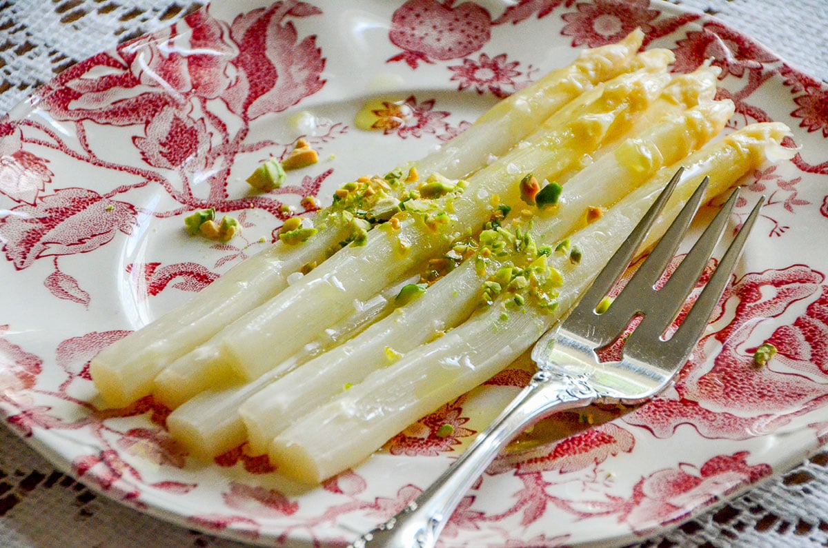 White asparagus on a pink plate with pistachios and a silver fork on the side