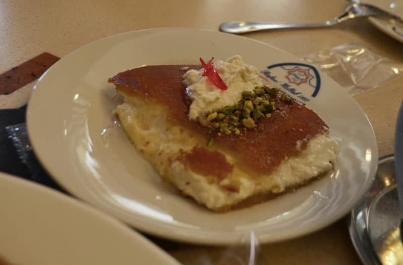 A square slice of knafeh pastry with golden top, a dollop of white cream and chopped pistachios on a white plate that reads Hallab 