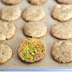 Sesame cookies on a parchment lined baking sheet with one turned over with pistachios on it