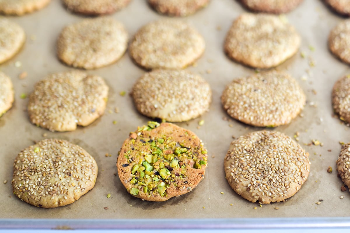 Sesame cookies on a parchment lined baking sheet with one turned over with pistachios on it