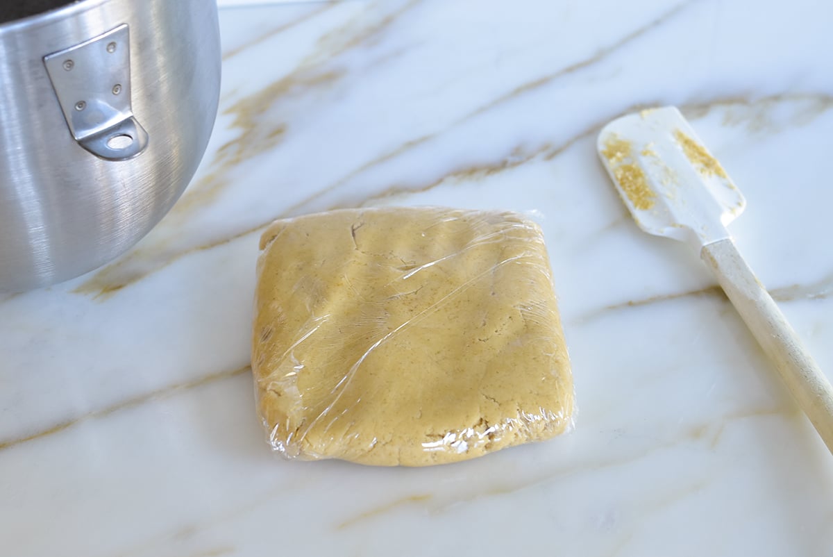 A square block of dough wrapped in plastic wrap with a rubber spatula on the side