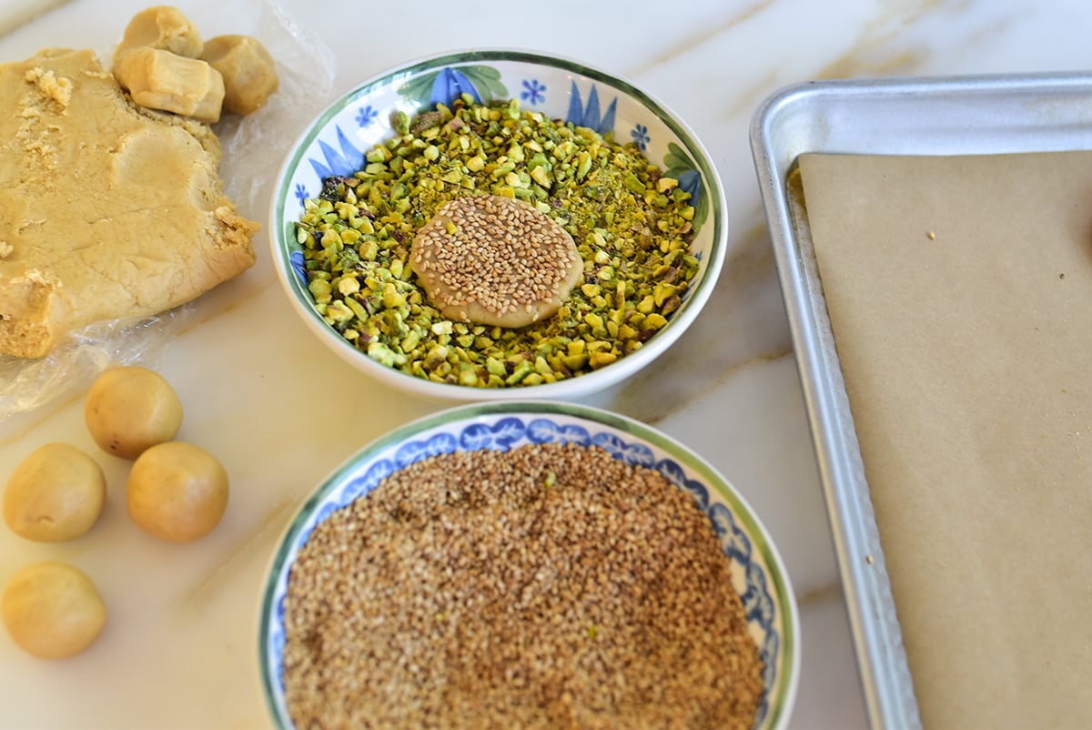 A flat disc of cookie dough coated with sesame seeds dipped in a bowl of chopped pistachios