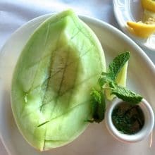 Ripe honeydew melon with a cup of fresh mint syrup and lemon wedges, on a white plate, Maureen Abood.com