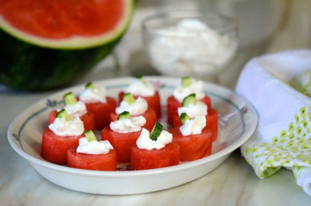 Watermelon bites with labneh and cucumber on a plate, sourrounded by watermelon and a green-trimmed towel, Maureen abood