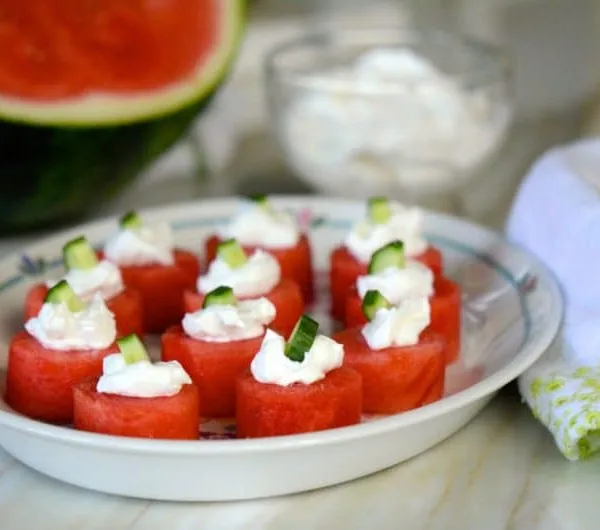 Watermelon bites with labneh and cucumber on a plate, sourrounded by watermelon and a green-trimmed towel, Maureen abood
