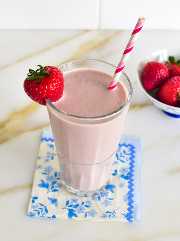 A pink smoothie in a tall glass with a strawberry on the side and a straw in the glass
