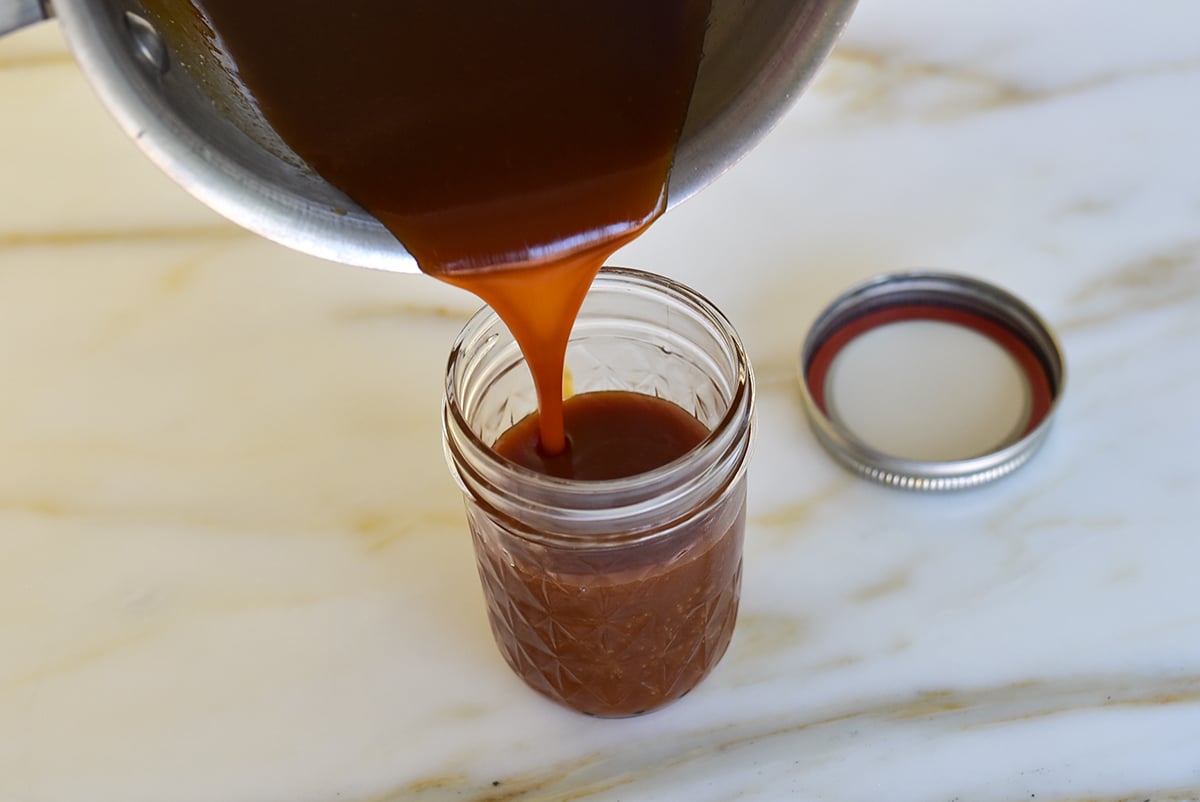 Homemade caramel sauce pouring from pot into a glass jar on a marble counter