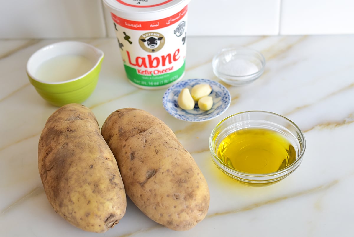 Ingredients for mashed potatoes with two potatoes, olive oil, labneh, garlic cloves, salt and milk in small bowls