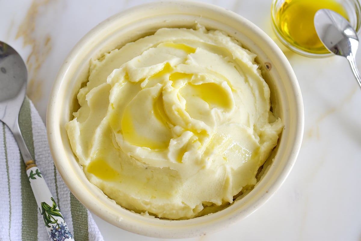 Olive oil mashed potatoes in a white dish with a spoon and a little bowl of olive oil on the side