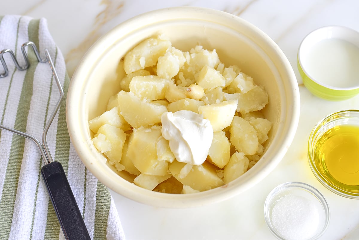 Cooked potatoes in a white dish with labneh on top and a masher on the side