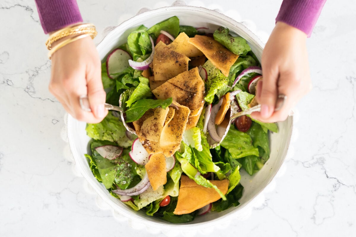 Fattoush salad in a white bowl with two hands stirring it with spoons