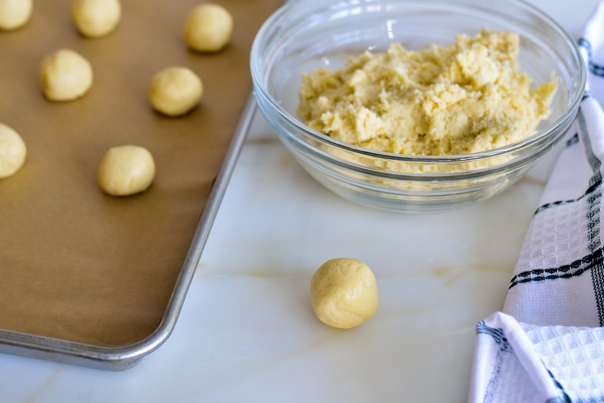 A dough ball beside a sheet of dough balls with a bowl of dough for thumbprint cookies