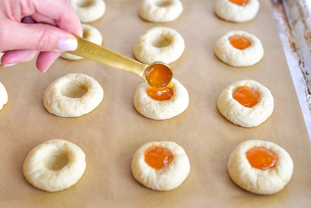 A spoon dropping apricot jam in the well of thumbprint cookies