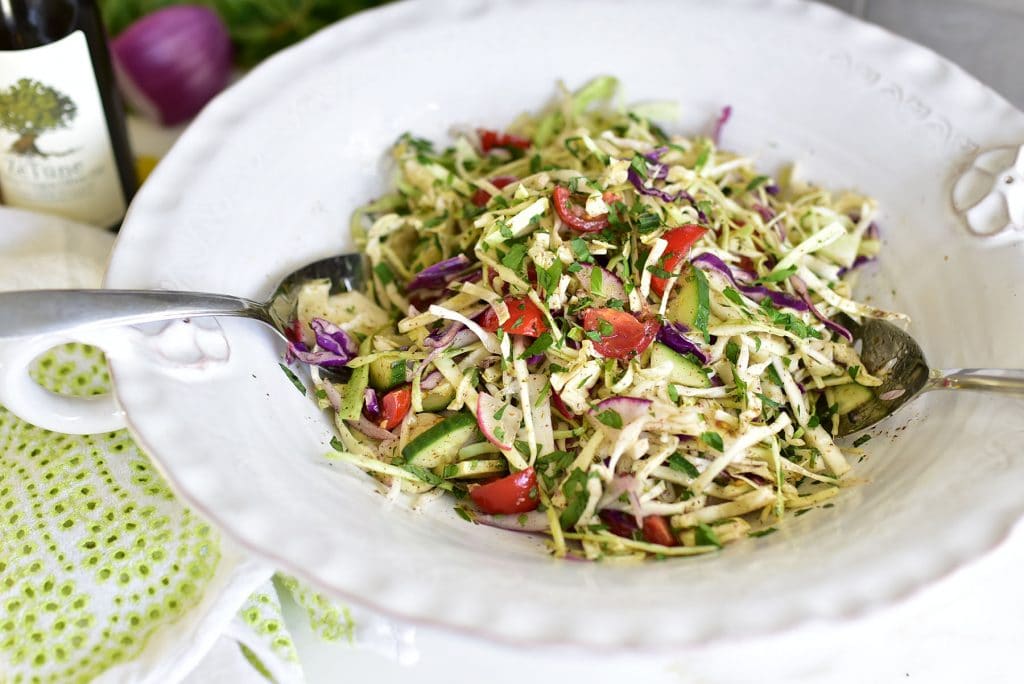 Cabbage slaw in a big white salad bowl with silver serving spoons