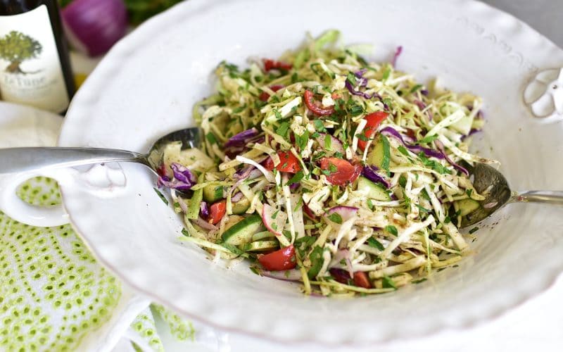 Cabbage slaw in a big white salad bowl with silver serving spoons