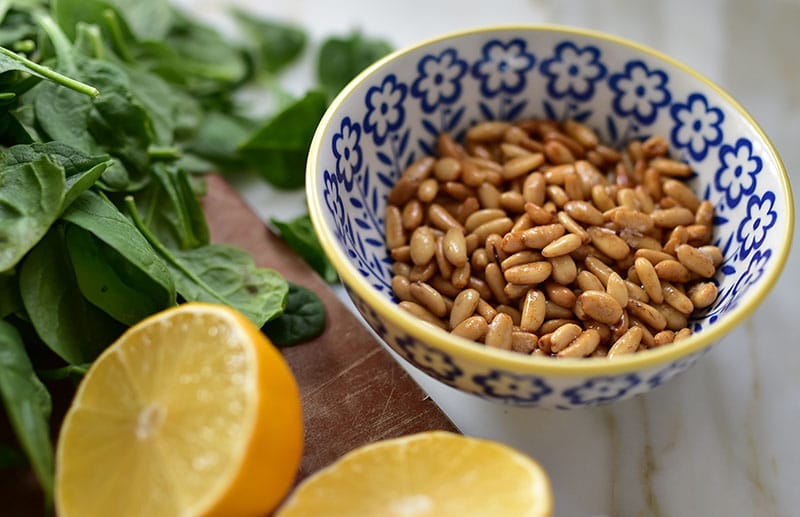 Toasted pine nuts in a small bowl next to a halved lemon.