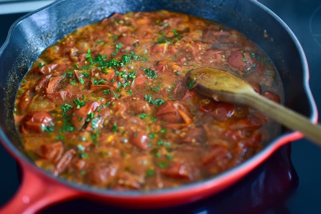 Tomatoes in a red saute pan with chopped fresh herbs, stirred with a wooden spoon