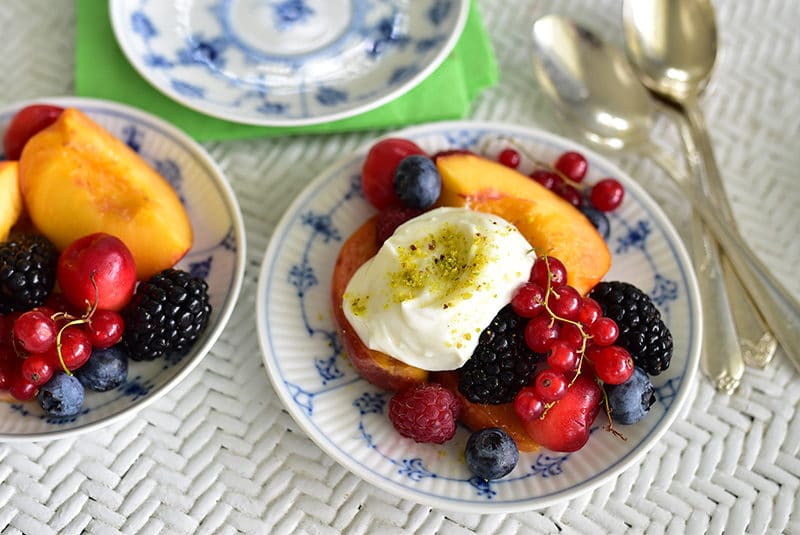 Fresh summer fruit with Lebanese ashta cream on a blue and white plate, surrounded by a wicker table and silver spoons from Maureen Abood