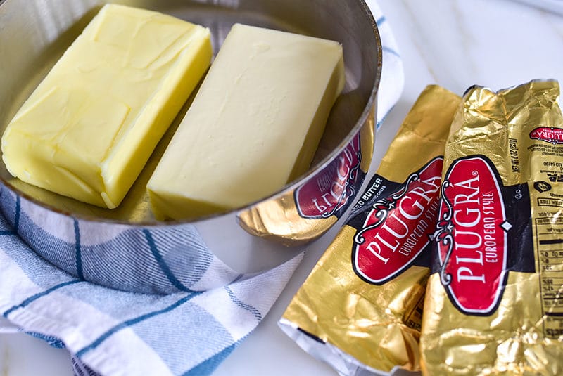 Plugra butter blocks in a pan on a blue and white towel with foil butter wrappers on the side