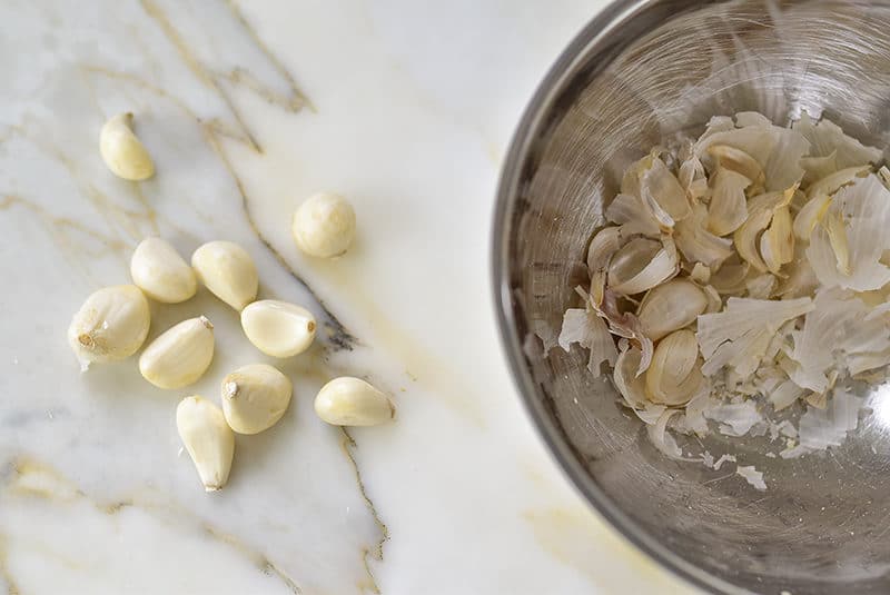 peeled garlic cloves next to a metal bowl full of garlic peels