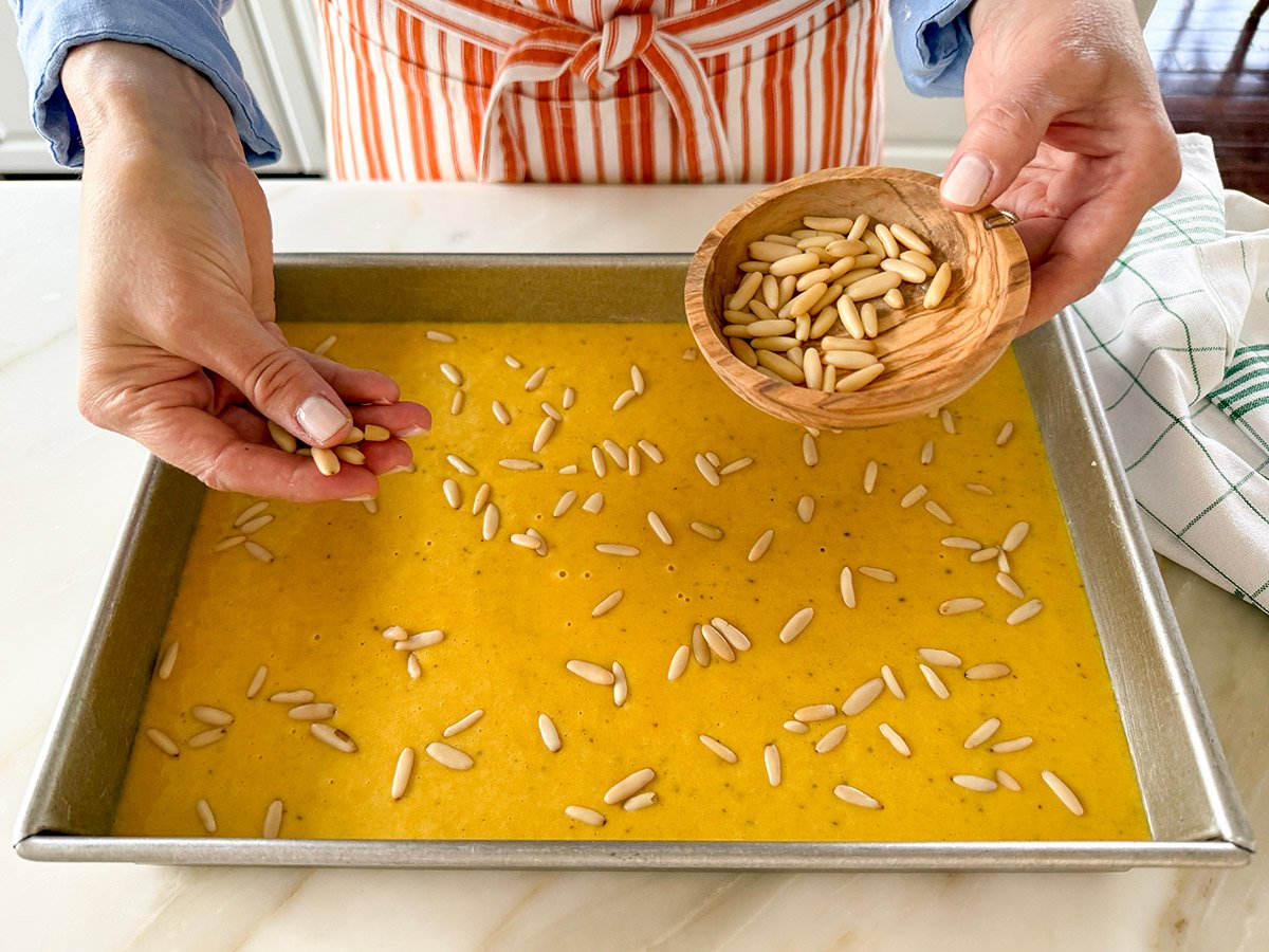Hands scattering pine nuts over sfouf cake batter in a pan