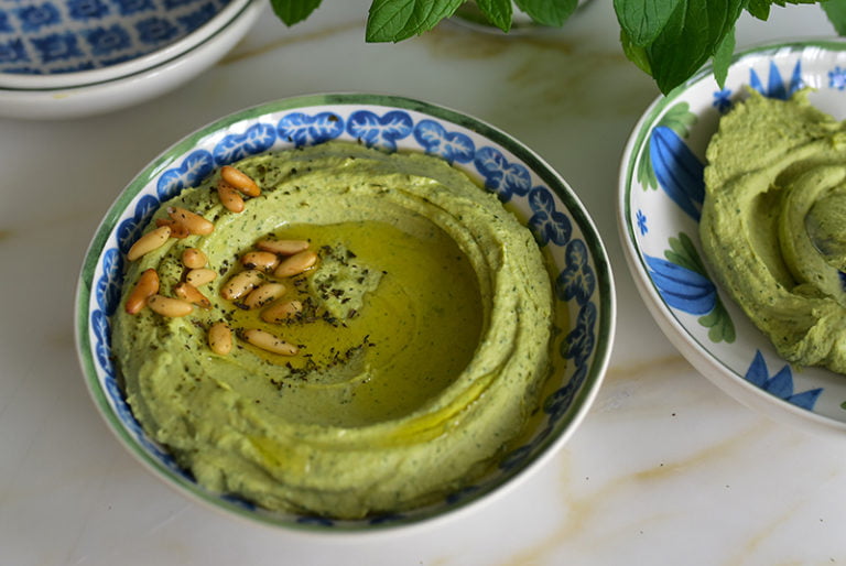 Green mint hummus swirled in a blue bowl with toasted pine nuts on top