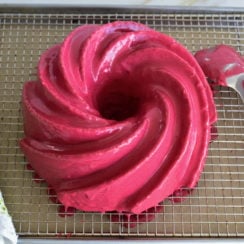 Pink glazed bundt cake on a rack over a sheet pan with a spoon on the side and green and white towel