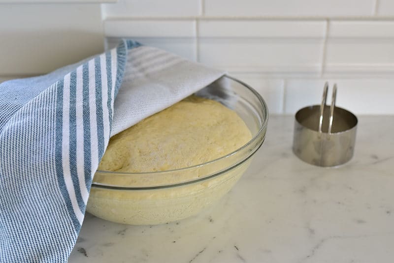 Dough in a glass bowl with a blue towel over the top and a round cutter on the side