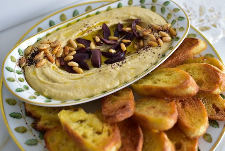 Hummus with olives, pine nuts, and crostini in an oval dish with crostini on the side