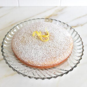 Olive oil cake on a glass platter with lemon zest and powdered sugar on top