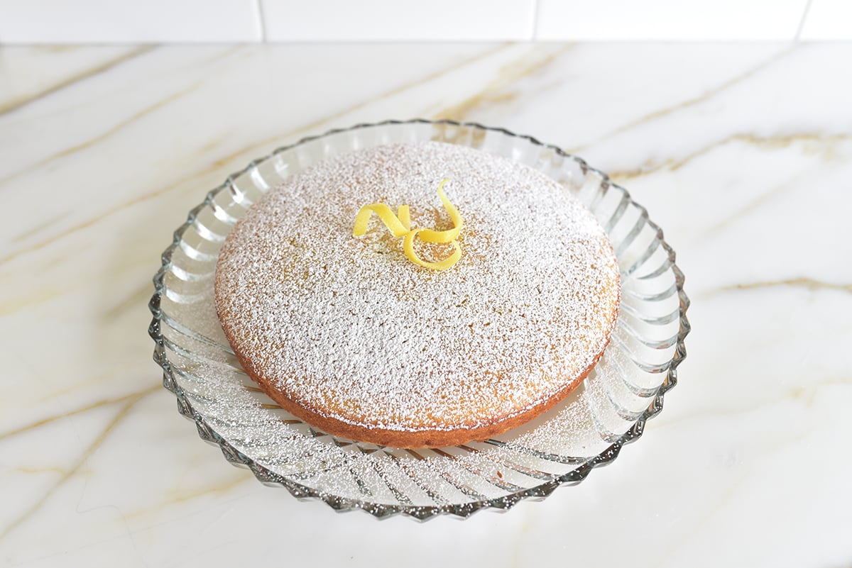 Olive oil cake on a glass platter with lemon zest and powdered sugar on top